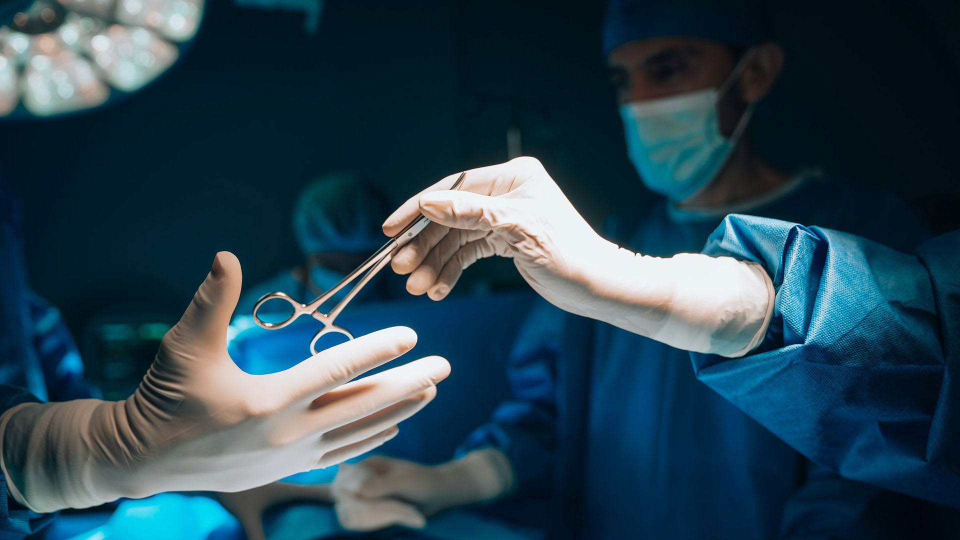 A medical team wearing PPE and passing a sterile surgical scissors to the surgeon.