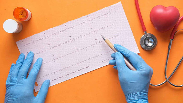 Hands donned with blue medical gloves above an ECG printout graph beside a pill box and a stethoscope.