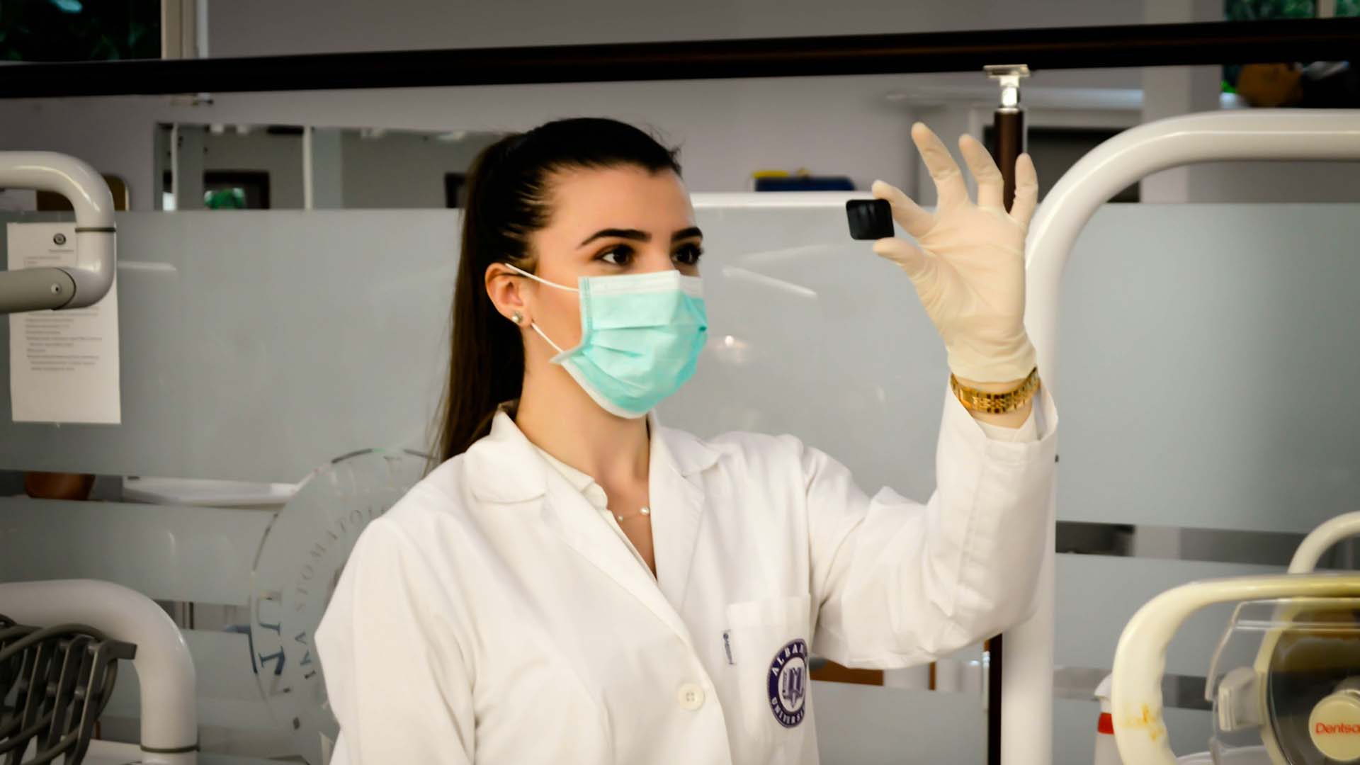 A dentist in lab coat and mask holds something up in front of her while wearing gloves.